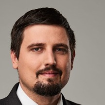 Portret mężczyzny z brodą, uśmiechniętego, na neutralnym szarym tle. Widoczny od ramion w górę, patrzy wprost w obiektyw. Profesjonalne oświetlenie.