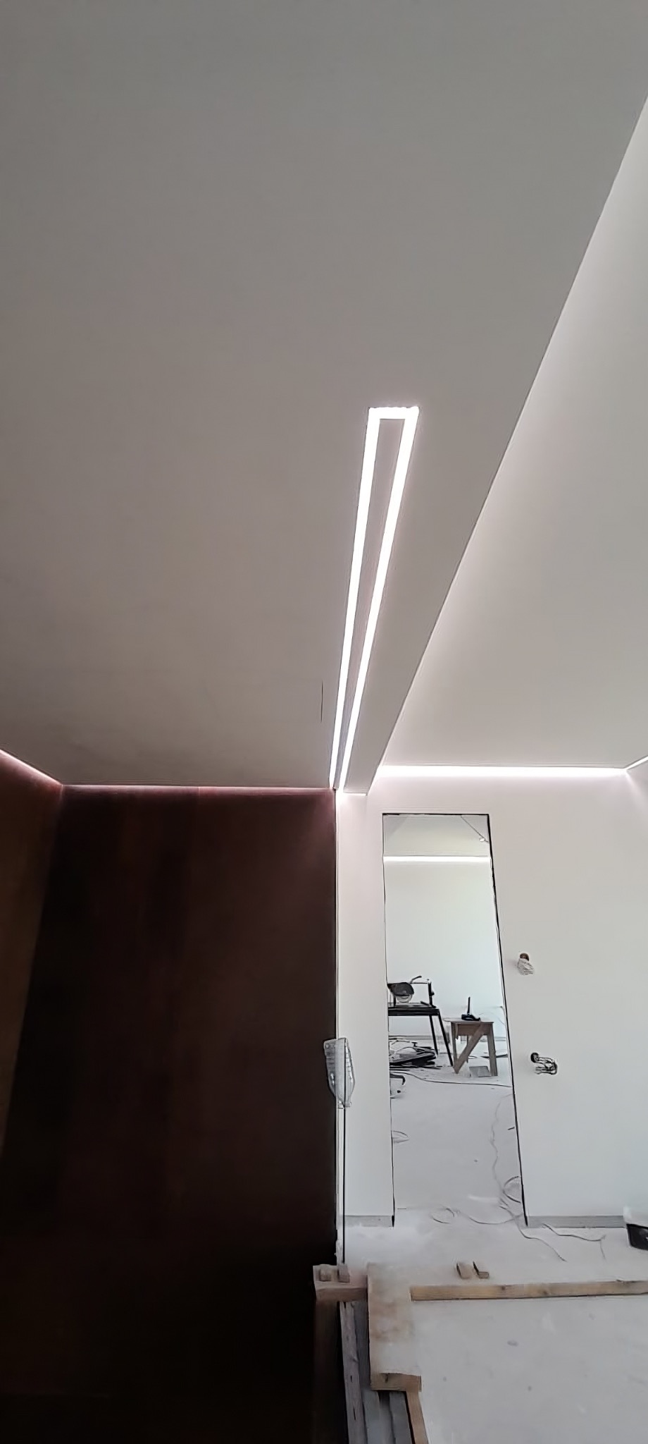 Nowoczesne oświetlenie sufitowe LED z profilami w minimalistycznym wnętrzu. Brązowa ściana i lustro w tle. Widoczne elementy wykończeniowe.