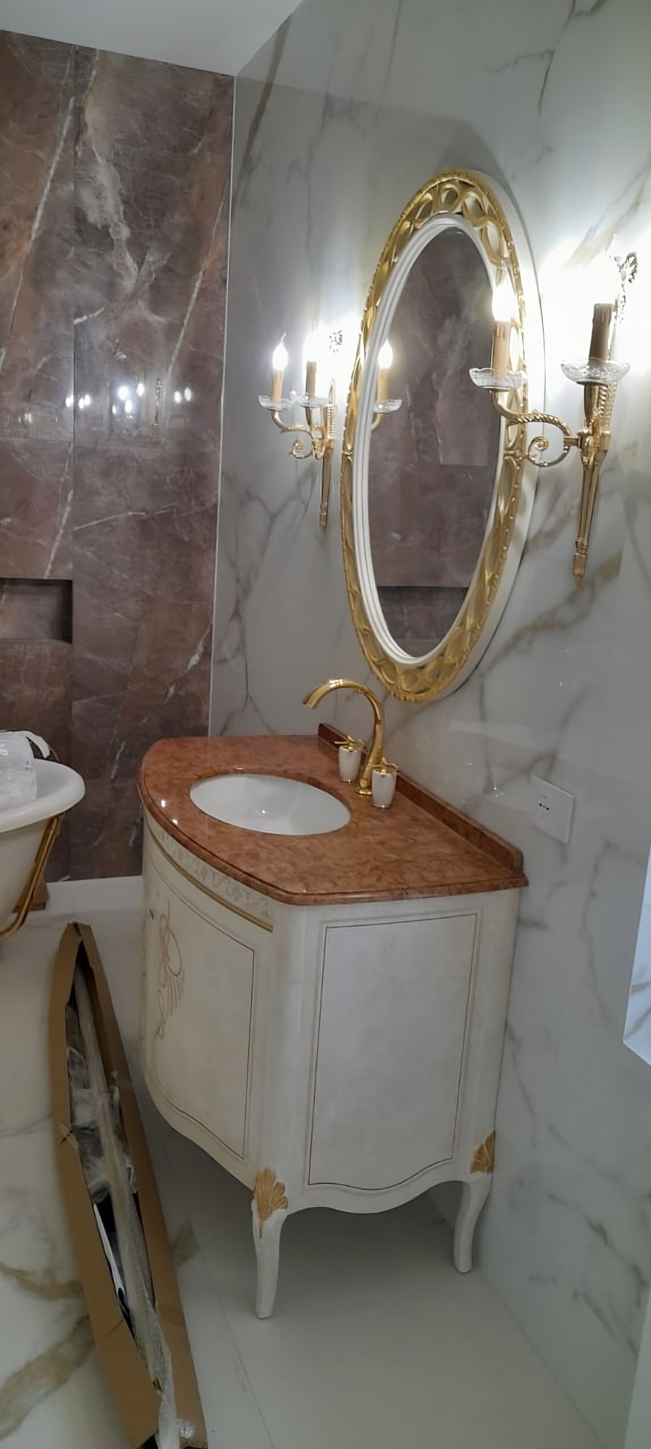 Elegancka łazienka w stylu glamour: marmurowe ściany, owalne lustro w złotej ramie, szafka umywalkowa z marmurowym blatem i złote akcenty.