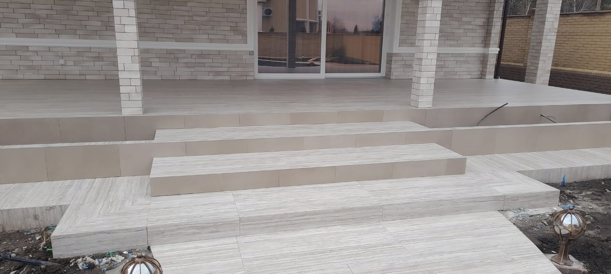 Wykończony taras z jasnymi płytkami imitującymi drewno, prowadzący do przeszklonych drzwi. Widoczne schody i kolumny wykończone tą samą płytką. Tło stanowi budynek i ogrodzenie.