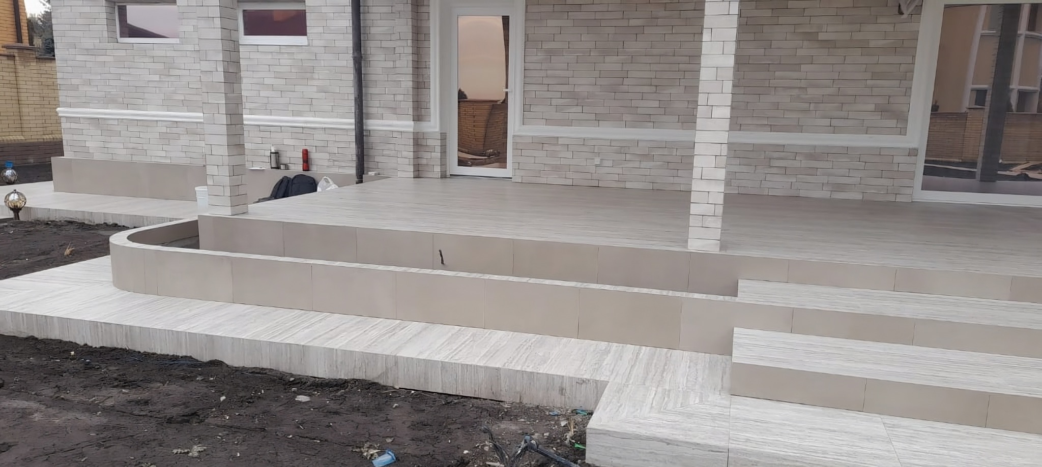 Wykończony taras z jasnoszarą płytką imitującą drewno. Schody wejściowe i obrzeża rabaty również wyłożone płytkami. Elewacja budynku z cegły w odcieniach szarości.