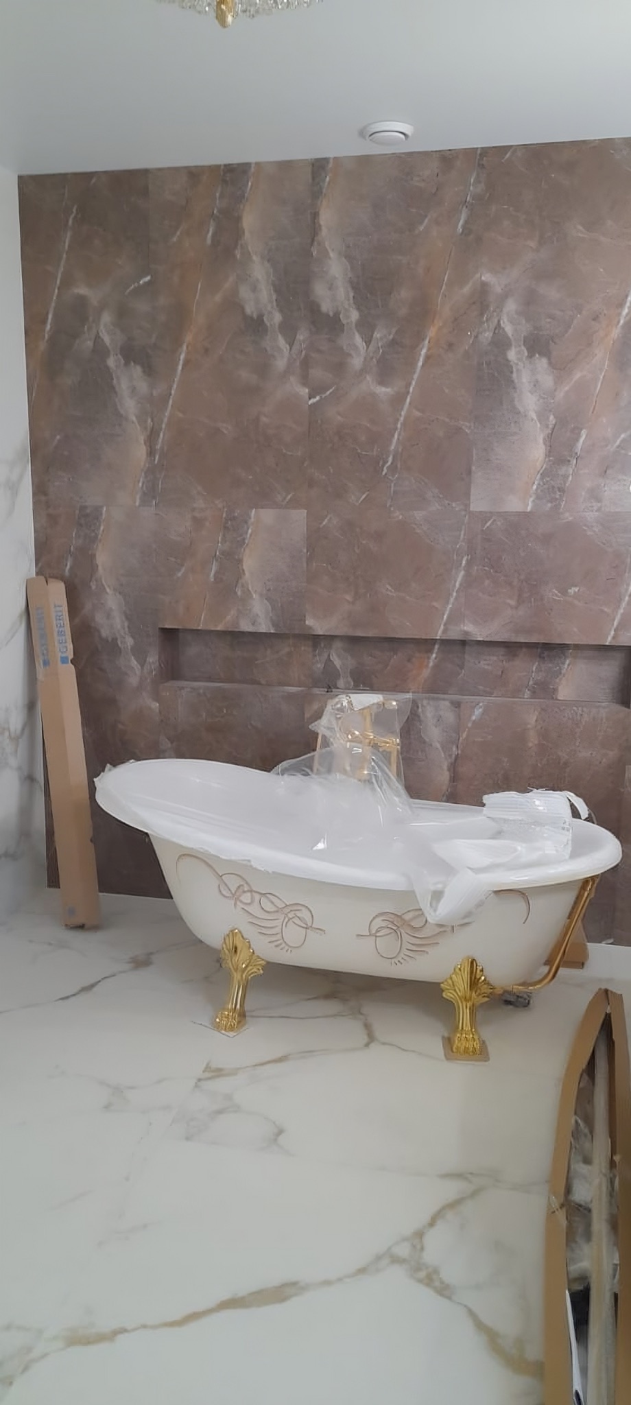 Elegancka, wolnostojąca wanna na złotych nóżkach, zabezpieczona folią, na tle ściany z płytek imitujących marmur. Widoczny fragment podłogi z białego marmuru.