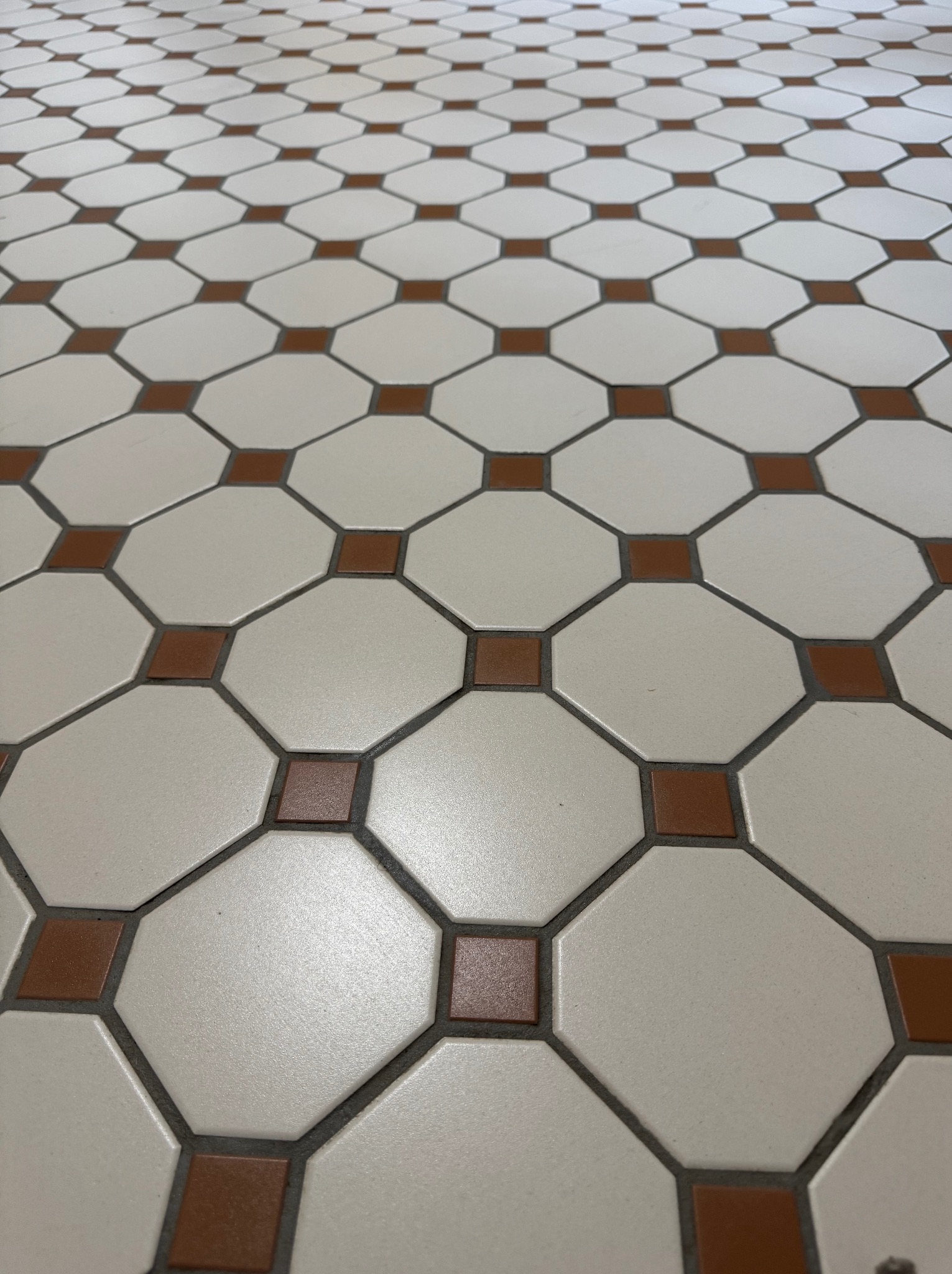 Ułożona podłoga z białych, ośmiokątnych płytek z brązowymi kwadratami w miejscach styku. Widok z perspektywy podłogi, fuga w kolorze szarym.