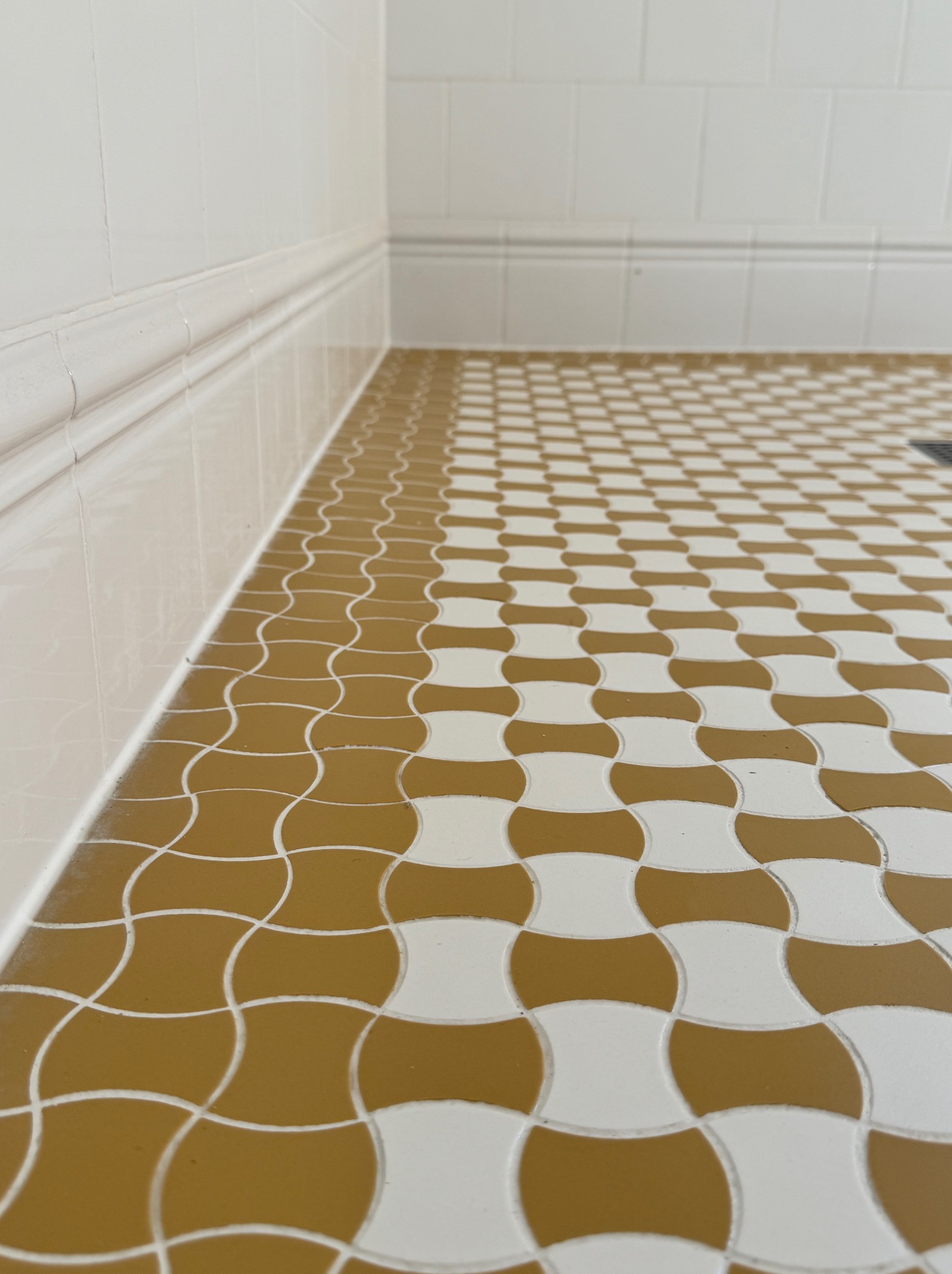 Beżowo-biała mozaika podłogowa w łazience, ułożona w charakterystyczny wzór. Widoczne białe płytki ścienne z dekoracyjnym wykończeniem. Perspektywa z dołu.