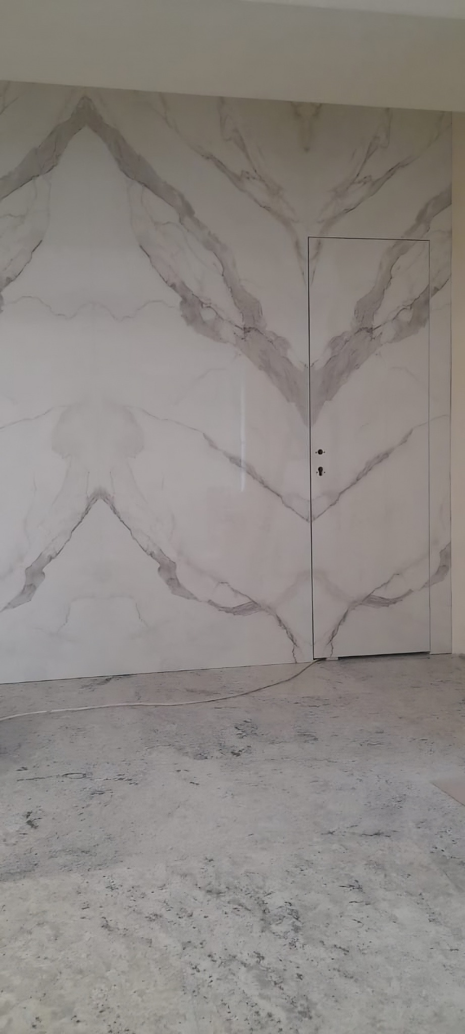 Ściana wyłożona płytami imitującymi marmur z ukrytymi drzwiami zamykanymi na zamek, widok na surową podłogę. Minimalistyczny design.