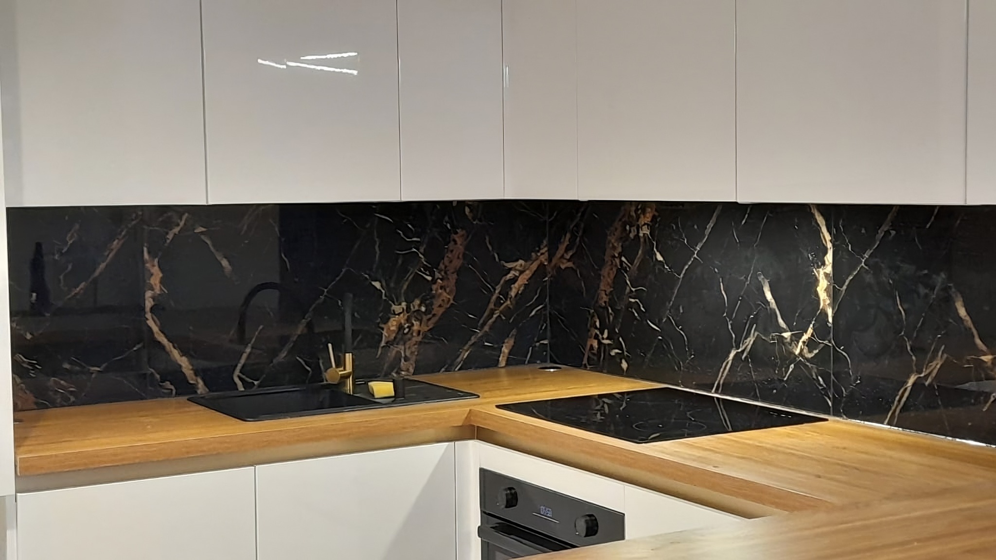 Nowoczesna kuchnia z czarnym panelem ściennym imitującym marmur, drewnianym blatem i białymi szafkami. Złota bateria przy czarnym zlewie i płyta indukcyjna.