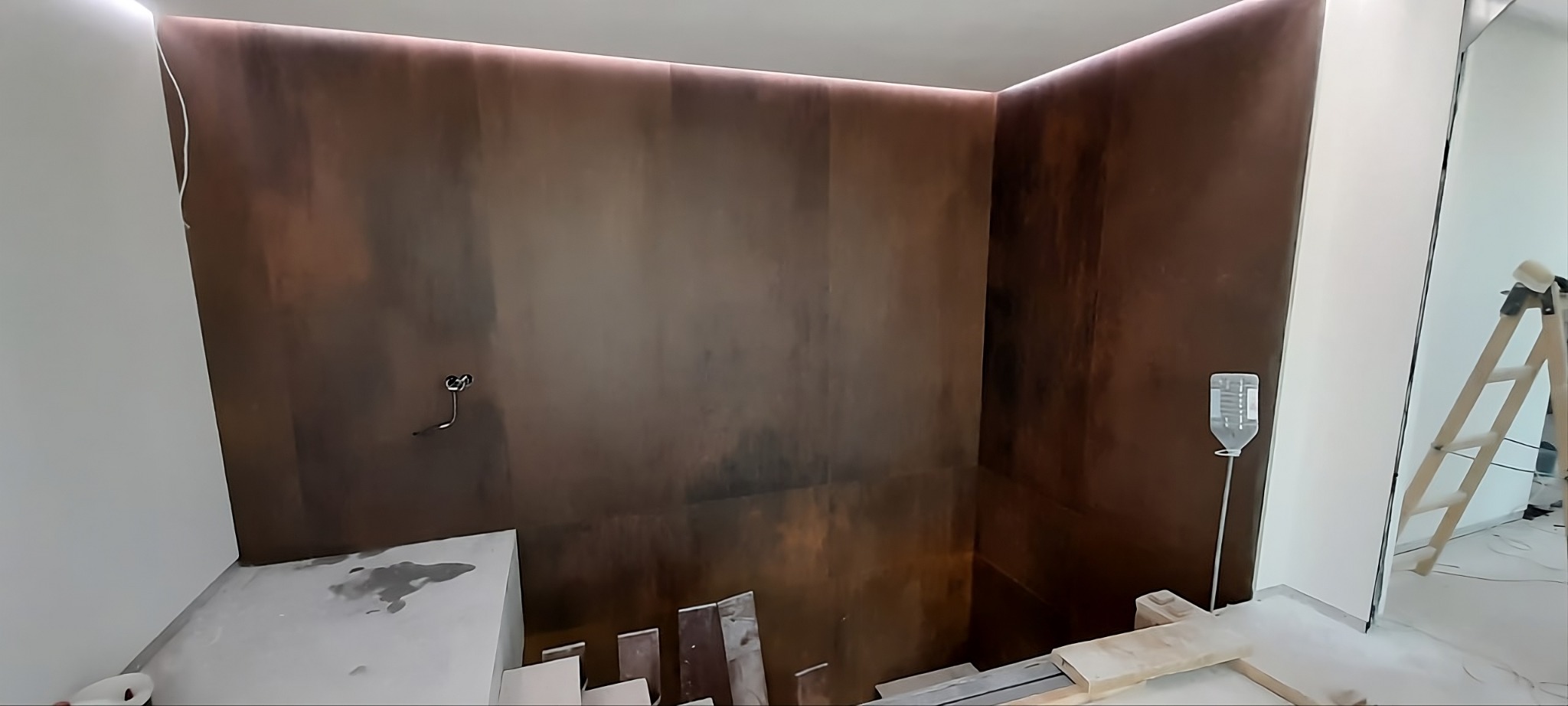 Nowoczesna łazienka w trakcie wykończenia: ściany z efektem rdzy, podświetlenie LED i drabina malarska. Widoczne materiały budowlane na podłodze.