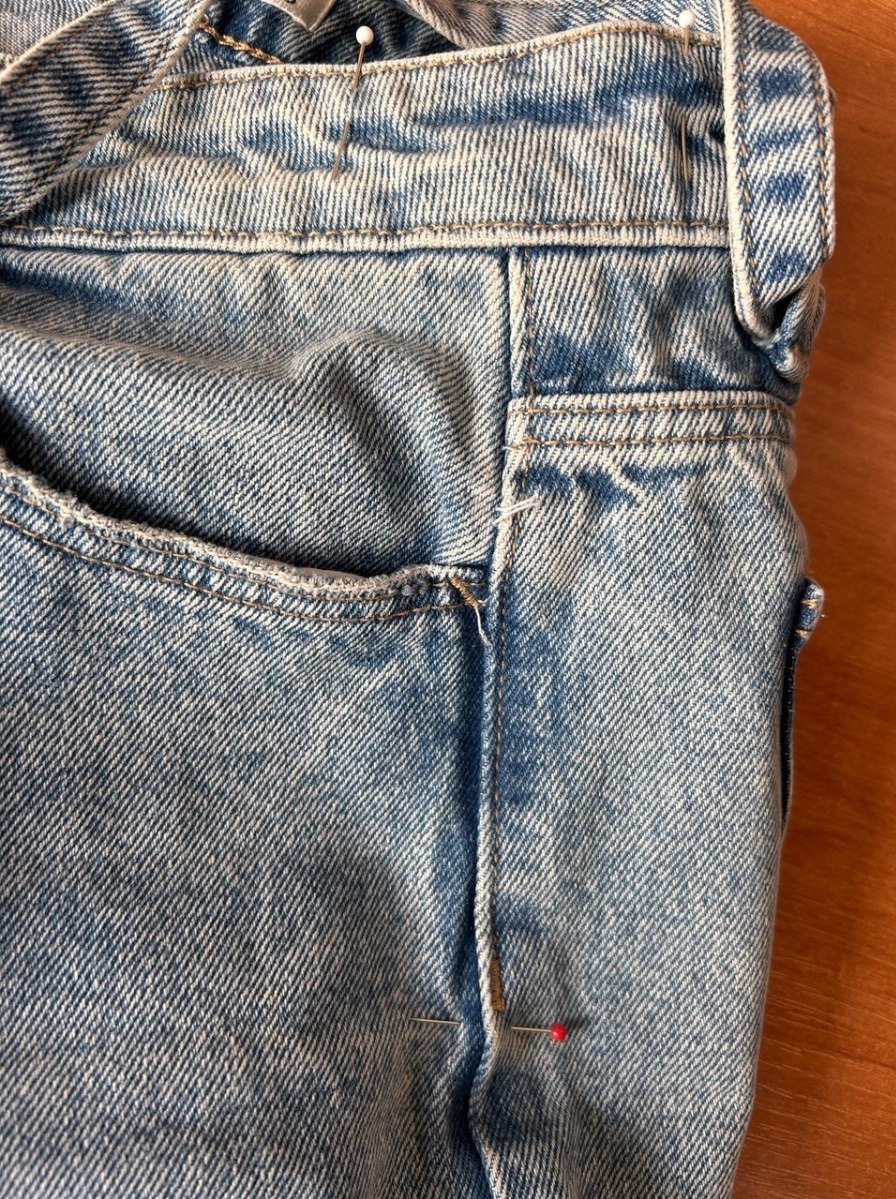 Fragment jasnych jeansów z widocznymi szpilkami krawieckimi, sugerujący proces dopasowywania lub poprawki. Detal kieszeni i szwów.