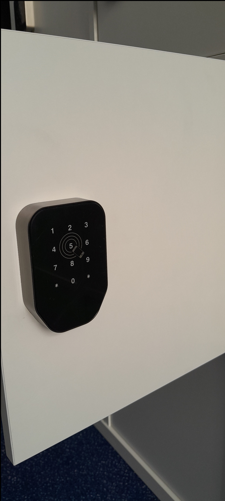 Zamontowany, czarny zamek szyfrowy z klawiaturą numeryczną na białych drzwiach szafki w biurze. Zamek z wyświetlaczem i przyciskami dotykowymi.