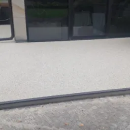 Jasna, beżowa nawierzchnia z żywicy epoksydowej na zewnątrz budynku, z widocznym obrzeżem i fragmentem elewacji. Nowoczesne wykończenie.