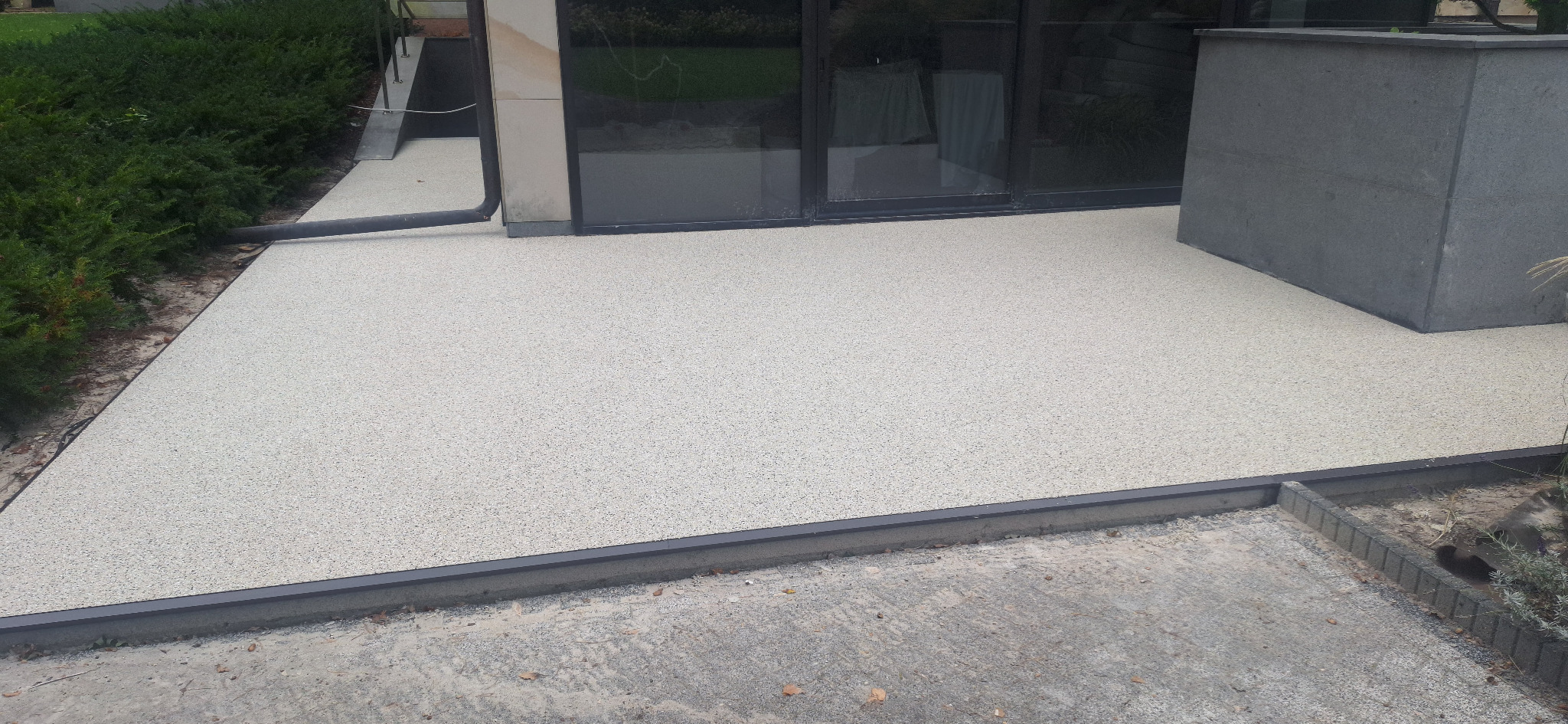 Jasna, beżowa nawierzchnia z żywicy epoksydowej na zewnątrz budynku, z widocznym obrzeżem i fragmentem elewacji. Nowoczesne wykończenie.