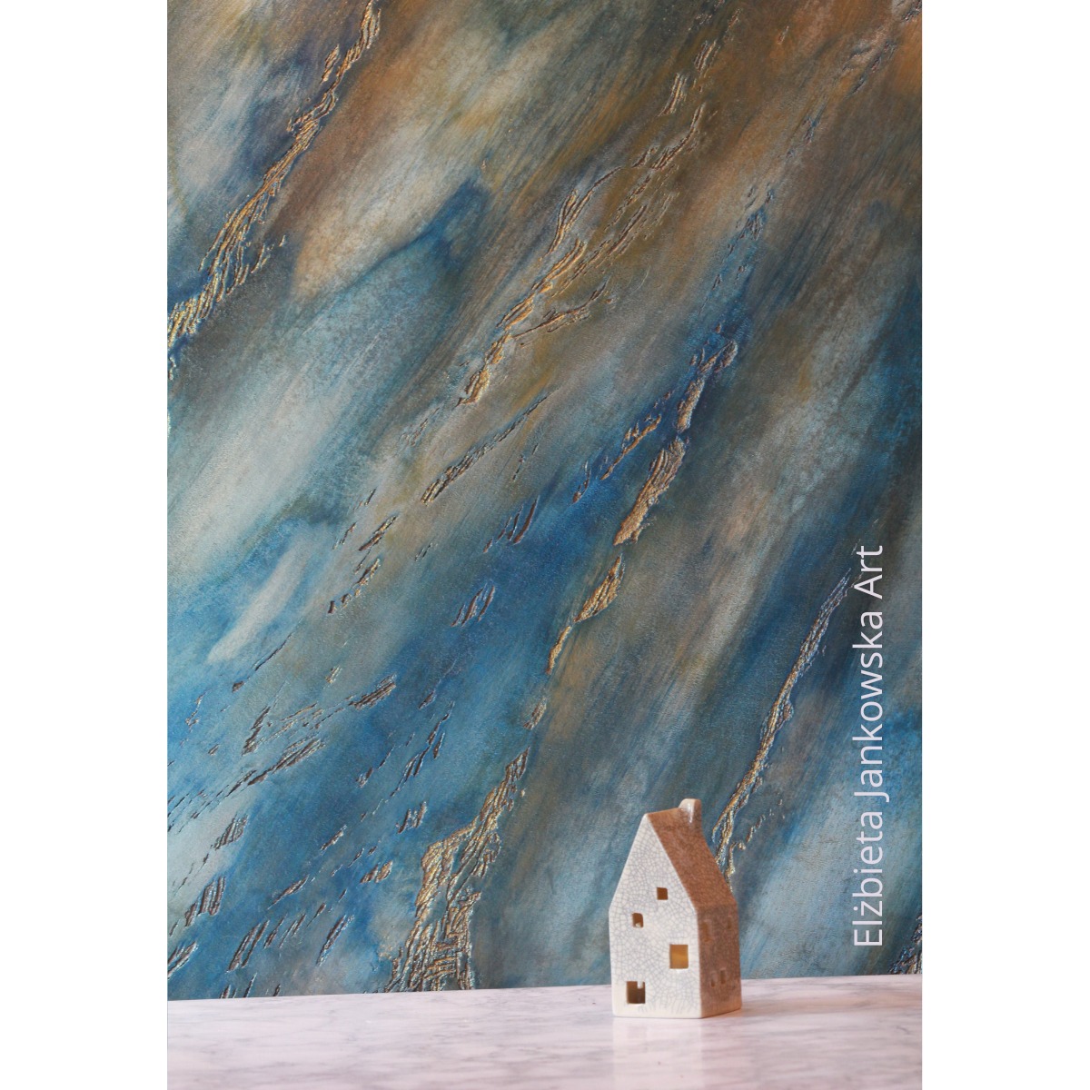 Dekoracyjna ściana z efektem labradorytu w odcieniach błękitu i złota, z delikatnym połyskiem. U dołu mała, ceramiczna figurka domu.