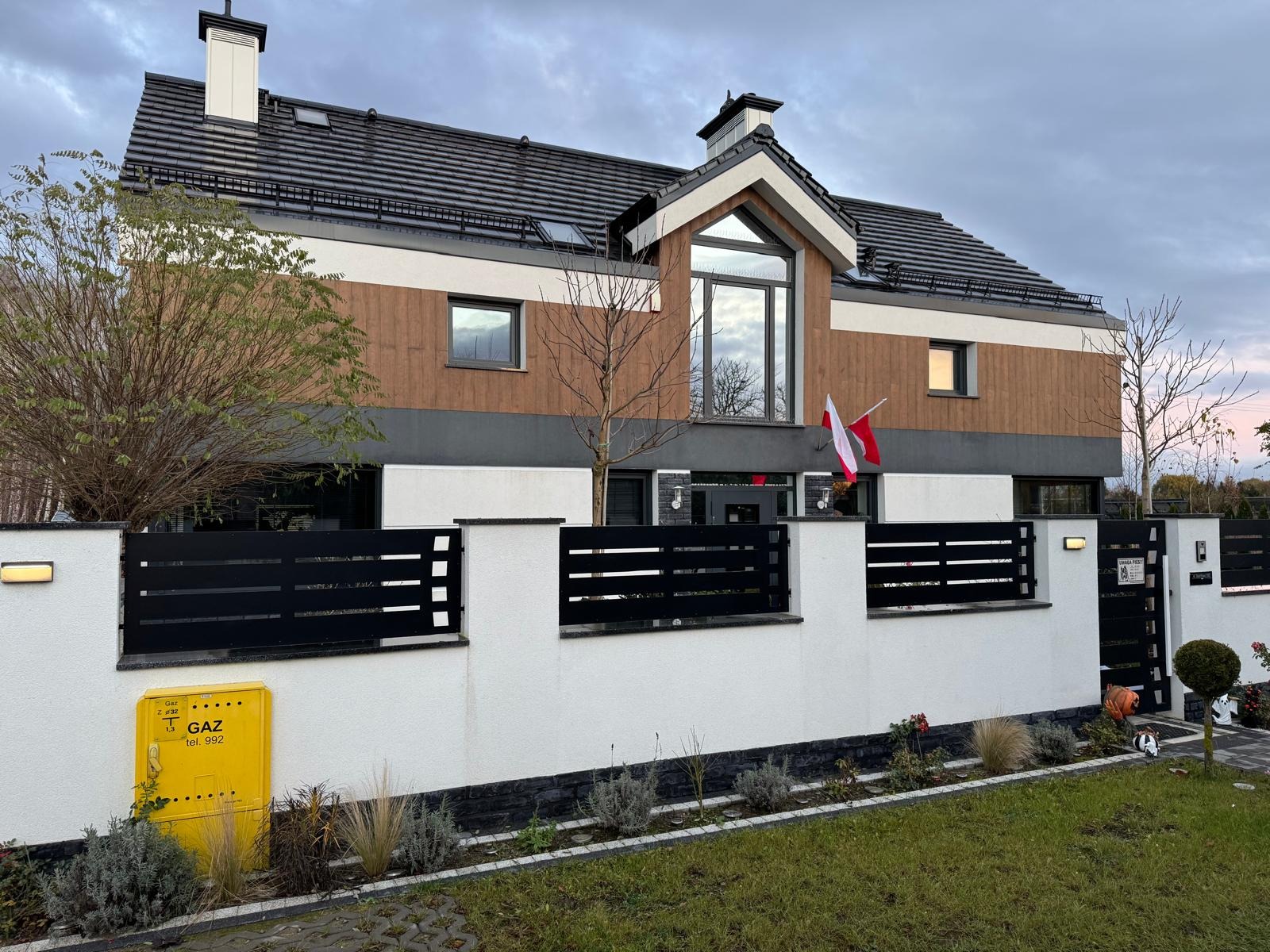 Elewacja domu jednorodzinnego z drewnianą okładziną i czarnym ogrodzeniem. Widoczna skrzynka gazowa i flaga Polski. Ujęcie z poziomu trawnika.