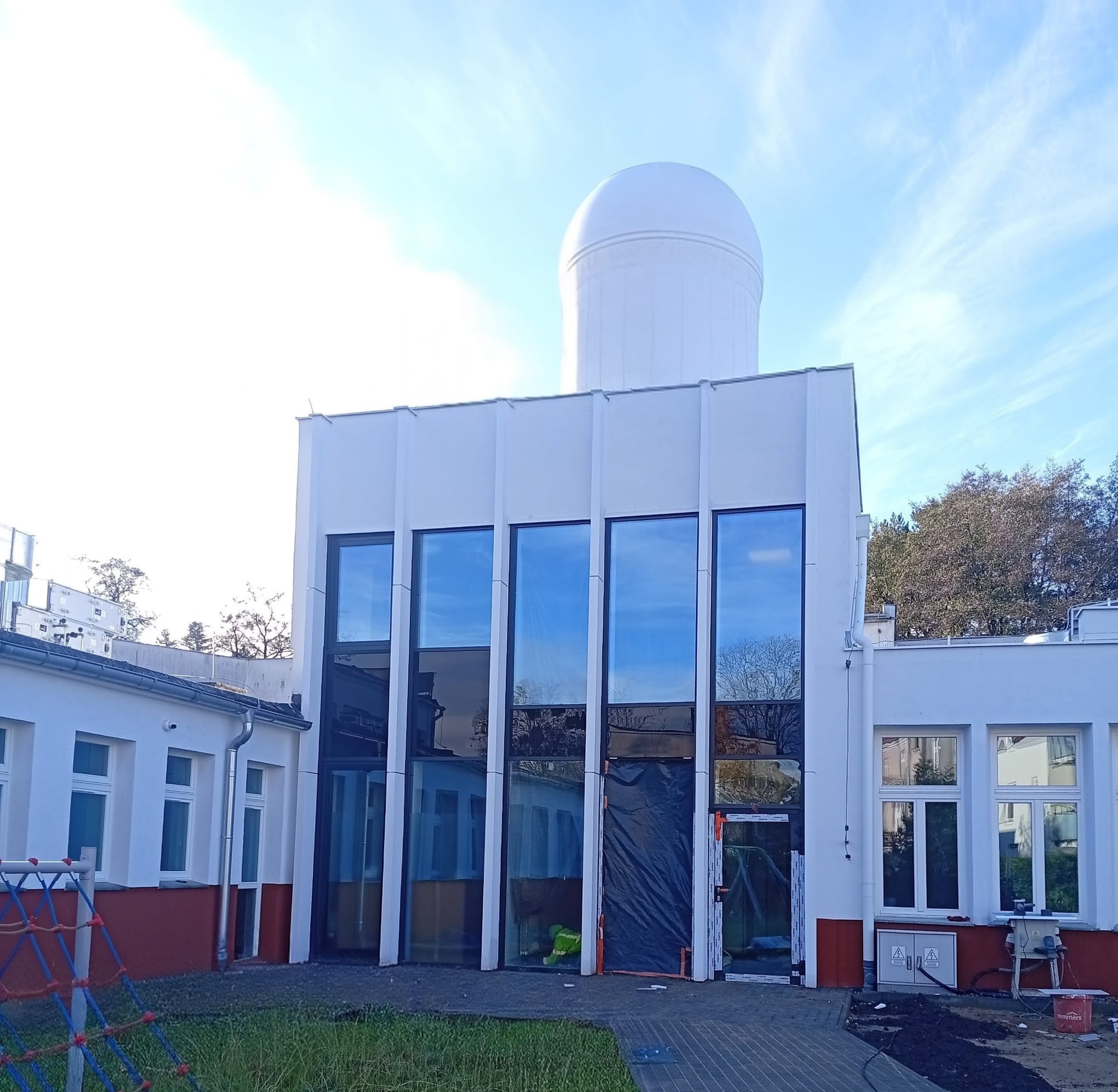 Biały budynek z kopułą obserwatorium, duże okna z odbiciem nieba, częściowo zakryte wejście, widoczne prace wykończeniowe na zewnątrz.