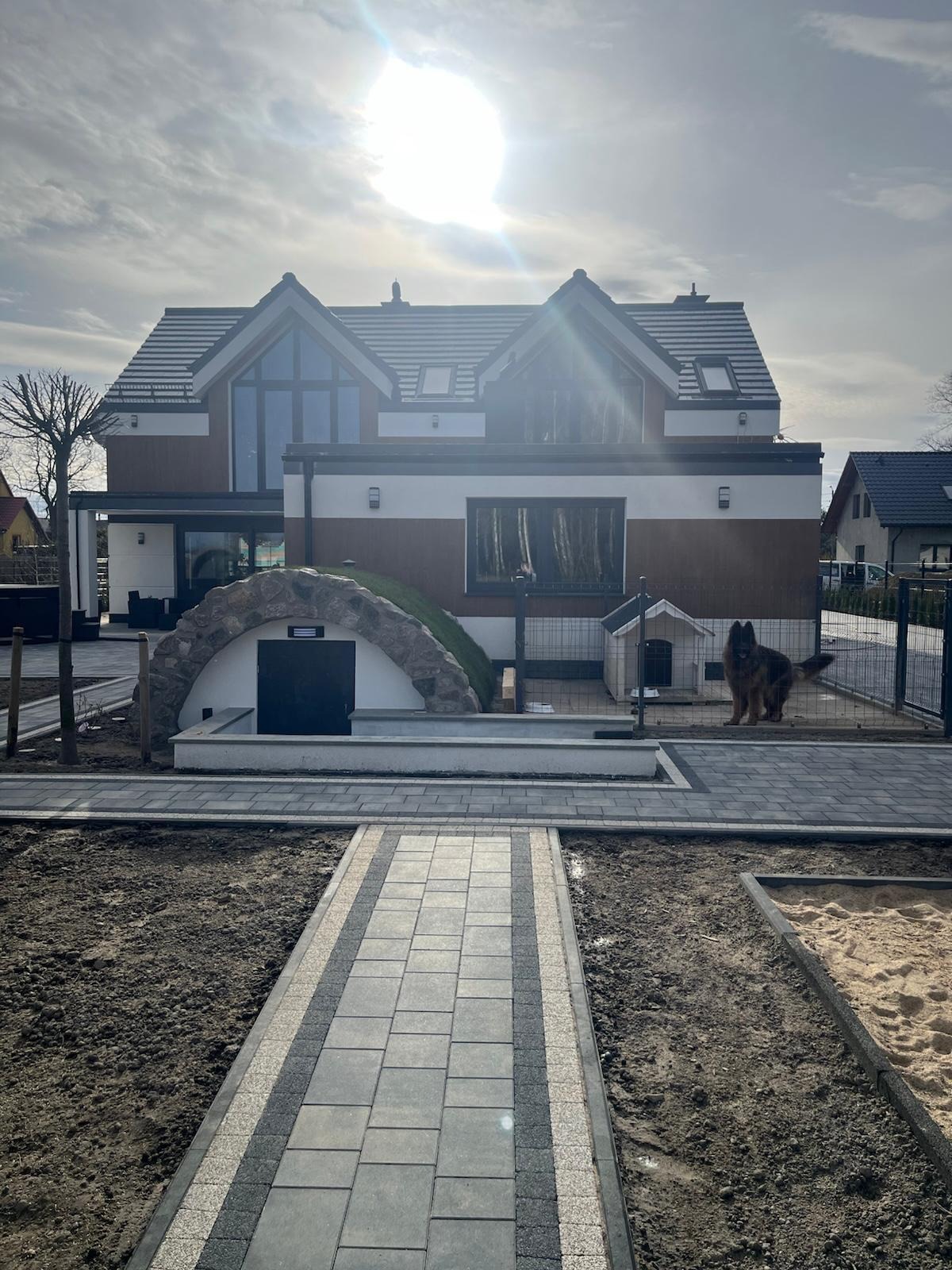 Nowoczesny dom z oryginalnym wejściem do piwnicy wykończonym kamieniem i zielenią. Ułożona kostka brukowa, widoczny pies rasy owczarek niemiecki.