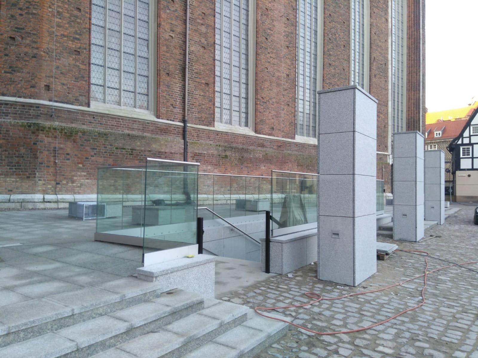 Zewnętrzne schody z szarego granitu prowadzą do przeszklonego wejścia. W tle ceglany budynek z wysokimi oknami i kamienne słupy. Widoczna kostka brukowa.
