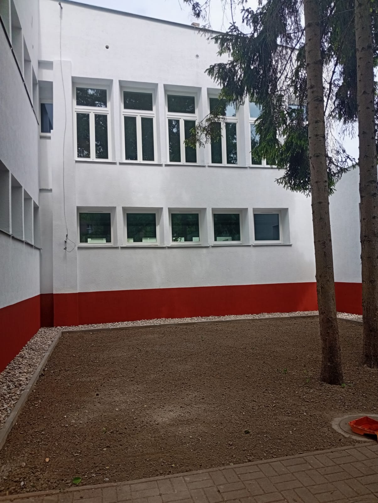 Biała fasada budynku z czerwonym pasem u dołu i rzędami okien, widok na przygotowany teren pod trawnik, obok drzewa iglaste.