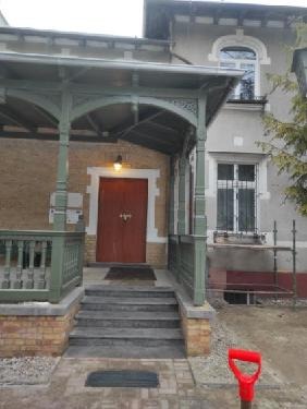 Wejście do budynku z drewnianą werandą w odcieniach zieleni i brązu, widoczne schody i czerwona łopata na pierwszym planie, elewacja w trakcie prac wykończeniowych.