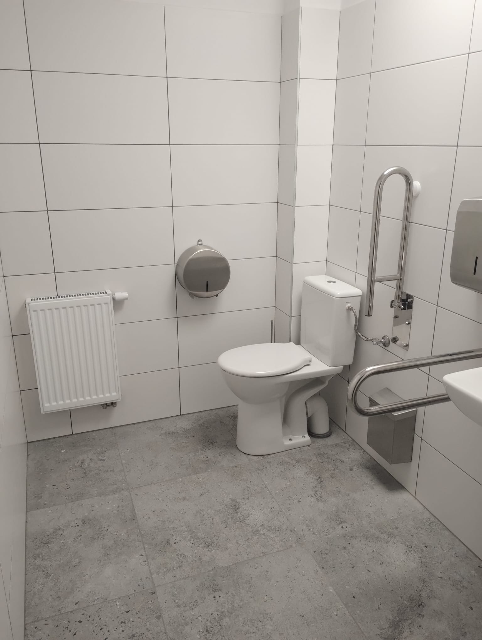 Jasna łazienka z białymi płytkami, toaletą i grzejnikiem. Uchwyty dla niepełnosprawnych zamontowane przy toalecie. Podłoga wyłożona szarymi płytkami.