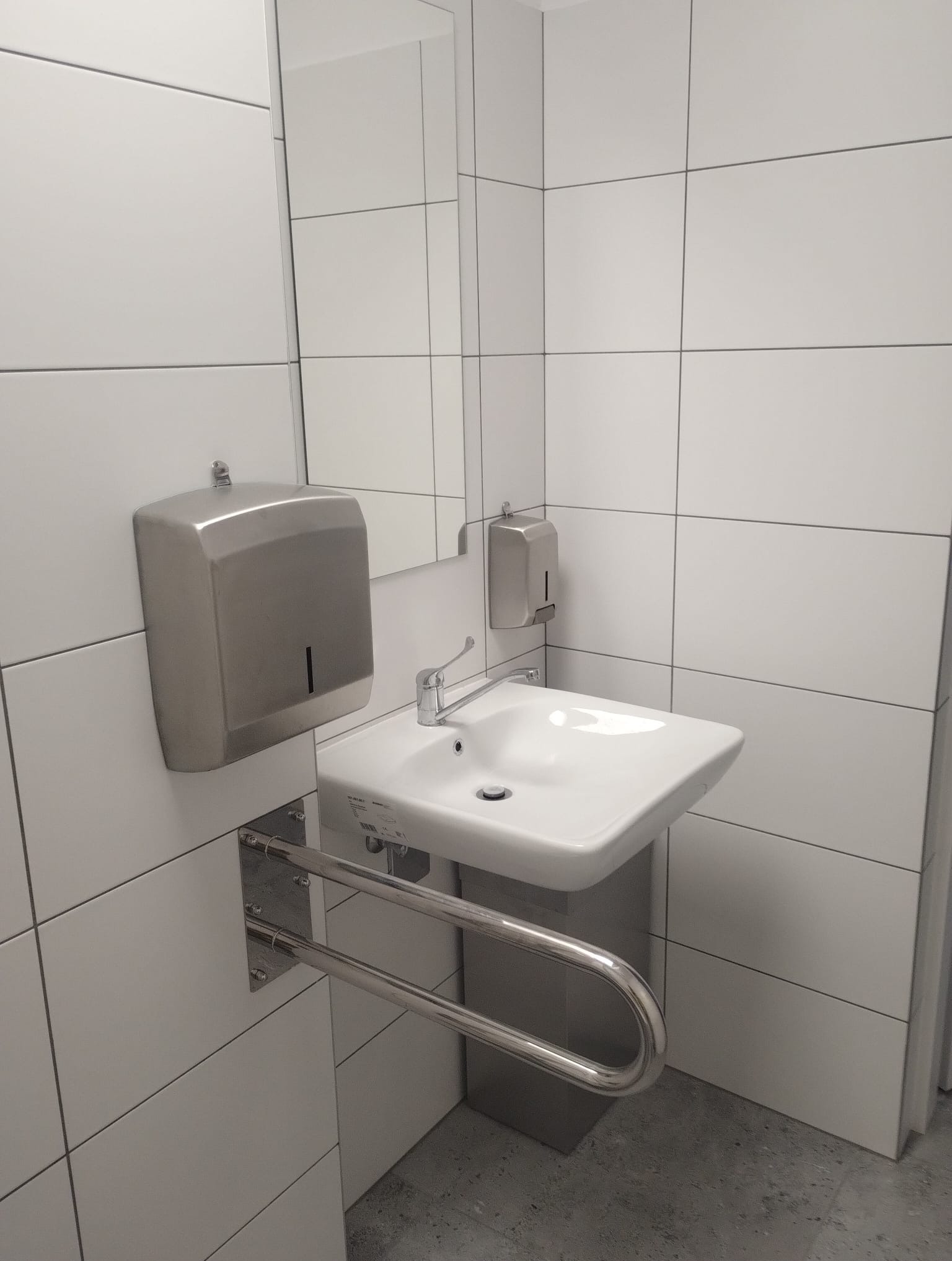 Biała umywalka z chromowaną baterią i uchwytem dla niepełnosprawnych na tle białych płytek w łazience. Widoczny dozownik mydła i lustro.