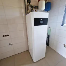 Biała pompa ciepła CTC z czarnym panelem, podłączona do instalacji z dwoma zbiornikami (biały i niebieski) na tle wykafelkowanej ściany i podłogi.