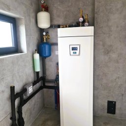 InstalCentrum Pompy Ciepła - Biała pompa ciepła w pomieszczeniu z szarymi płytkami, widoczne elementy instalacji: zbiorniki, rury. Minimalistyczny design, nowoczesne wykończenie.