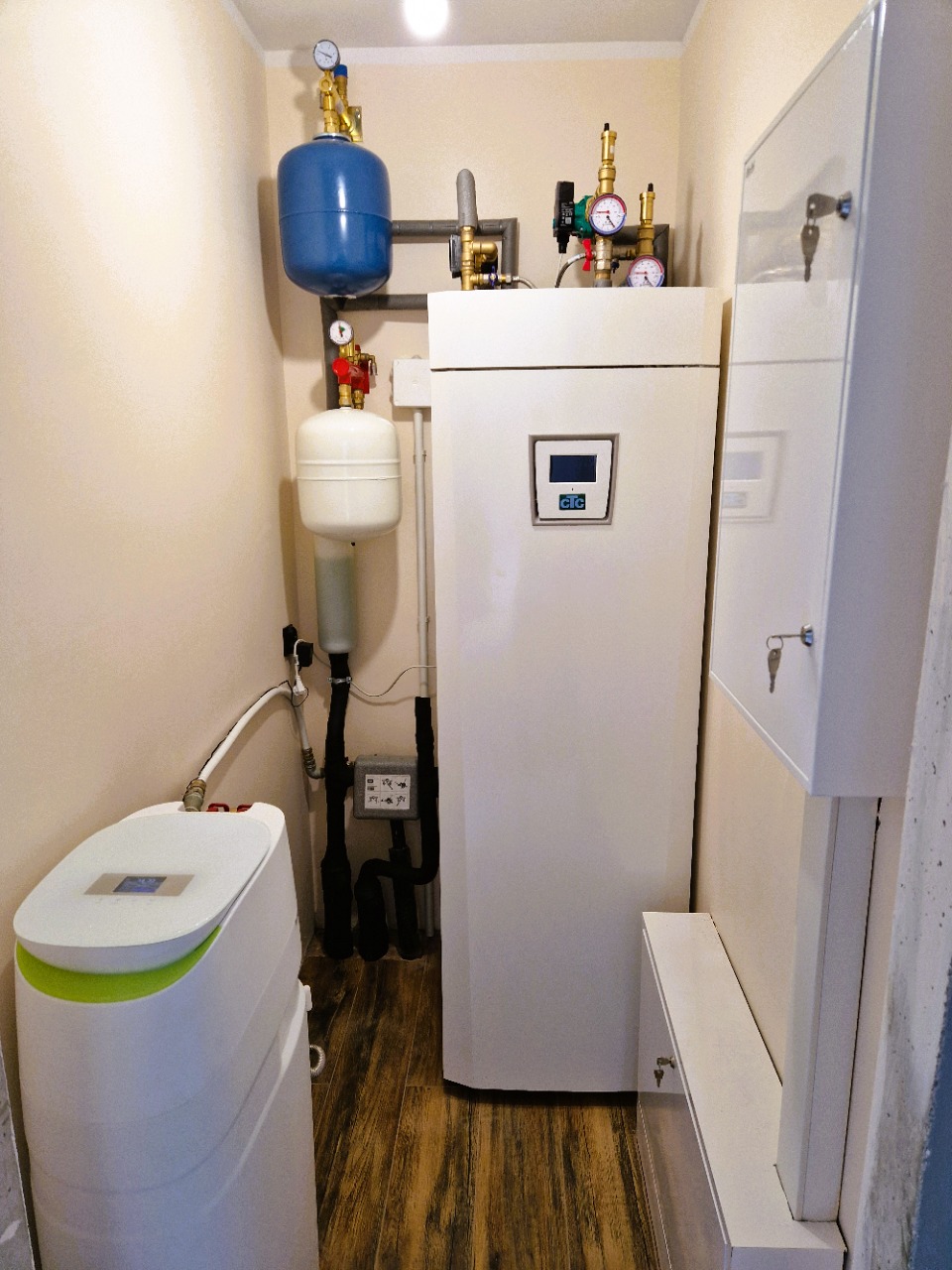 Kompaktowa kotłownia z białą pompą ciepła CTC, zbiornikiem hydroforowym i zmiękczaczem wody w narożnym pomieszczeniu z drewnopodobną podłogą.