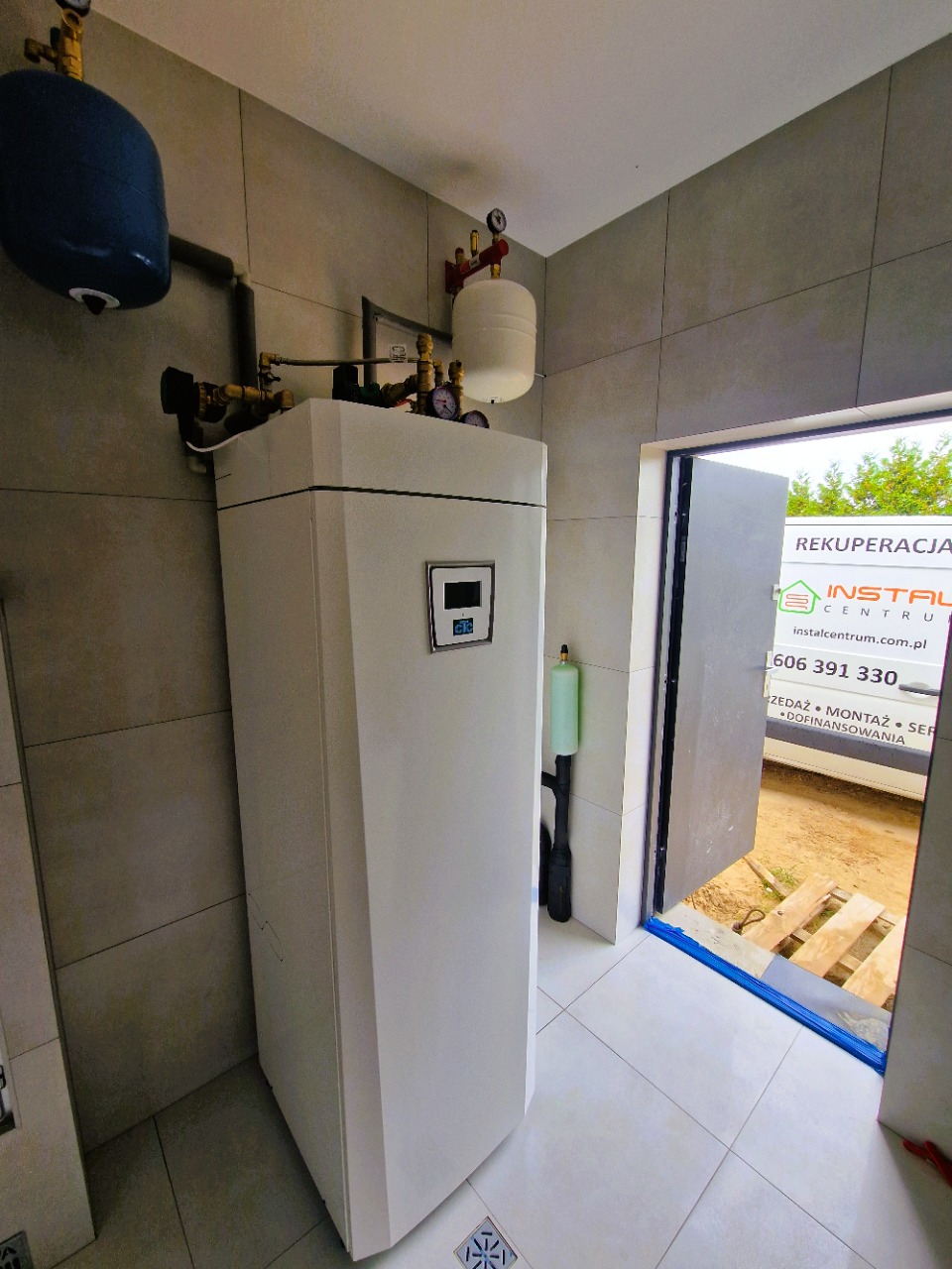 Biała pompa ciepła w kotłowni z szarymi płytkami w Lipnie. Widoczny zbiornik wyrównawczy i osprzęt. W tle otwarte drzwi i fragment samochodu dostawczego.