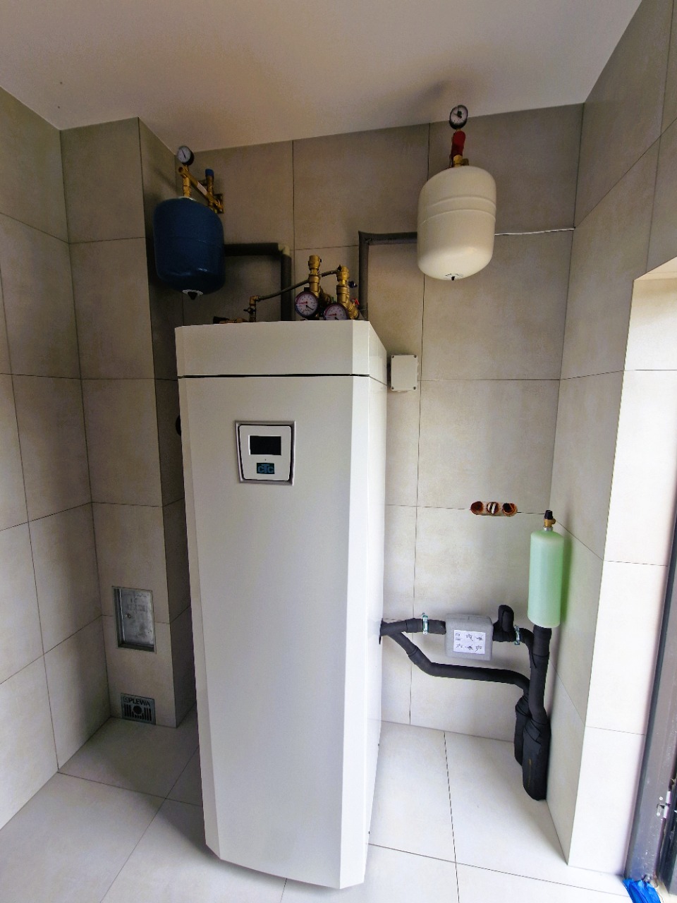 Biała pompa ciepła w kotłowni z beżowymi płytkami. Widoczne zbiorniki wyrównawcze, rury i manometry. Minimalistyczny design, czyste otoczenie.