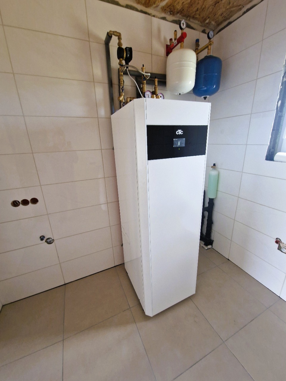 Biała pompa ciepła CTC z czarnym panelem, podłączona do instalacji z dwoma zbiornikami (biały i niebieski) na tle wykafelkowanej ściany i podłogi.