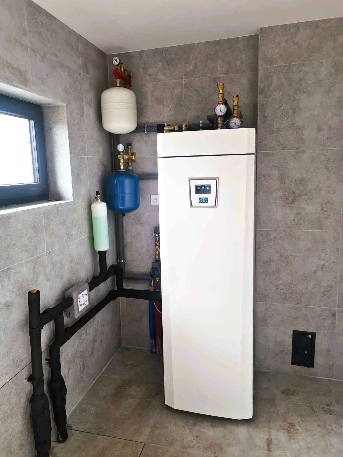 Biała pompa ciepła w pomieszczeniu z szarymi płytkami, widoczne elementy instalacji: zbiorniki, rury. Minimalistyczny design, nowoczesne wykończenie.