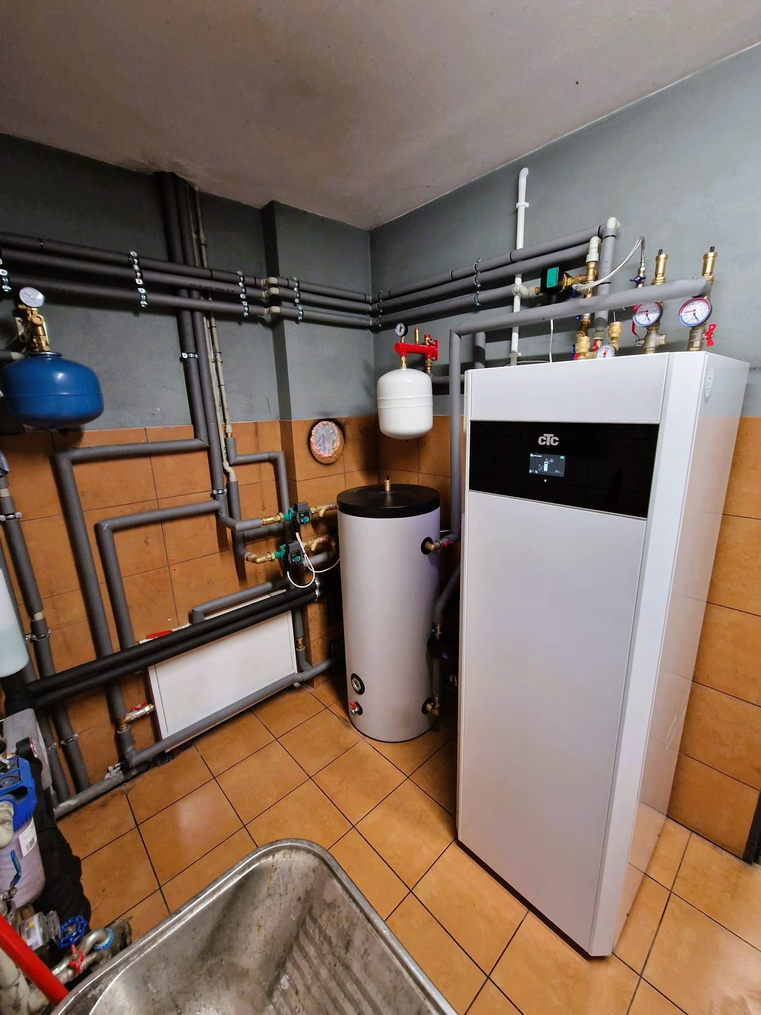 Biała pompa ciepła CTC z czarnym panelem, widoczna w kotłowni z instalacją rurową, zbiornikiem i osprzętem. Ujęcie z niskiego kąta, widoczne brązowe płytki na podłodze.