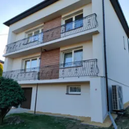 Dwupiętrowy dom z balkonami i elewacją w jasnym odcieniu. Kontrastują z nią czarne balustrady i rynny. Widoczny klimatyzator i fragment ogrodu z krzewem.