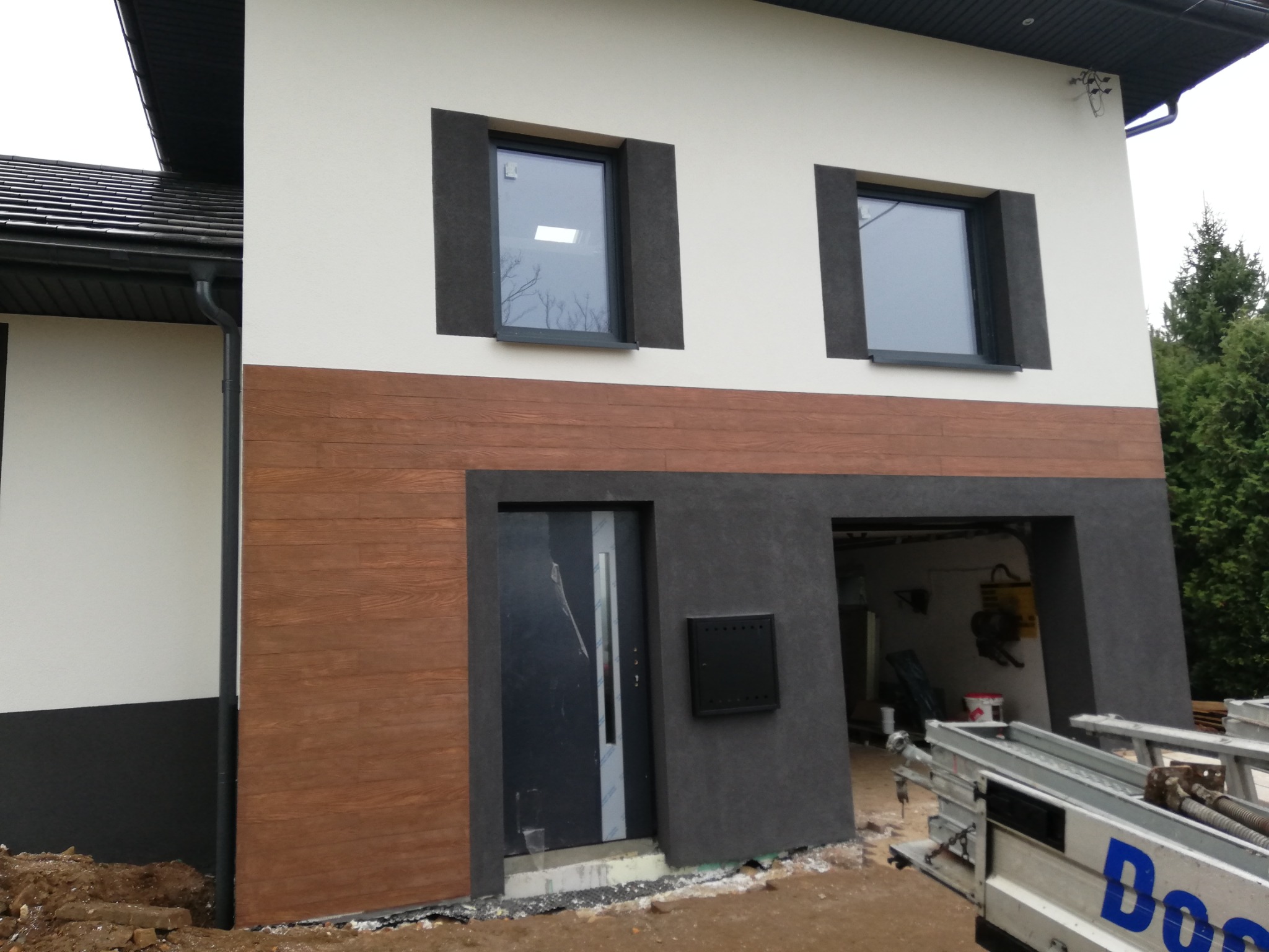 Wykończona elewacja domu z oknami w ciemnych obramowaniach, drewnianą okładziną i garażem. Widoczny fragment drabiny i teren wokół domu w trakcie prac.