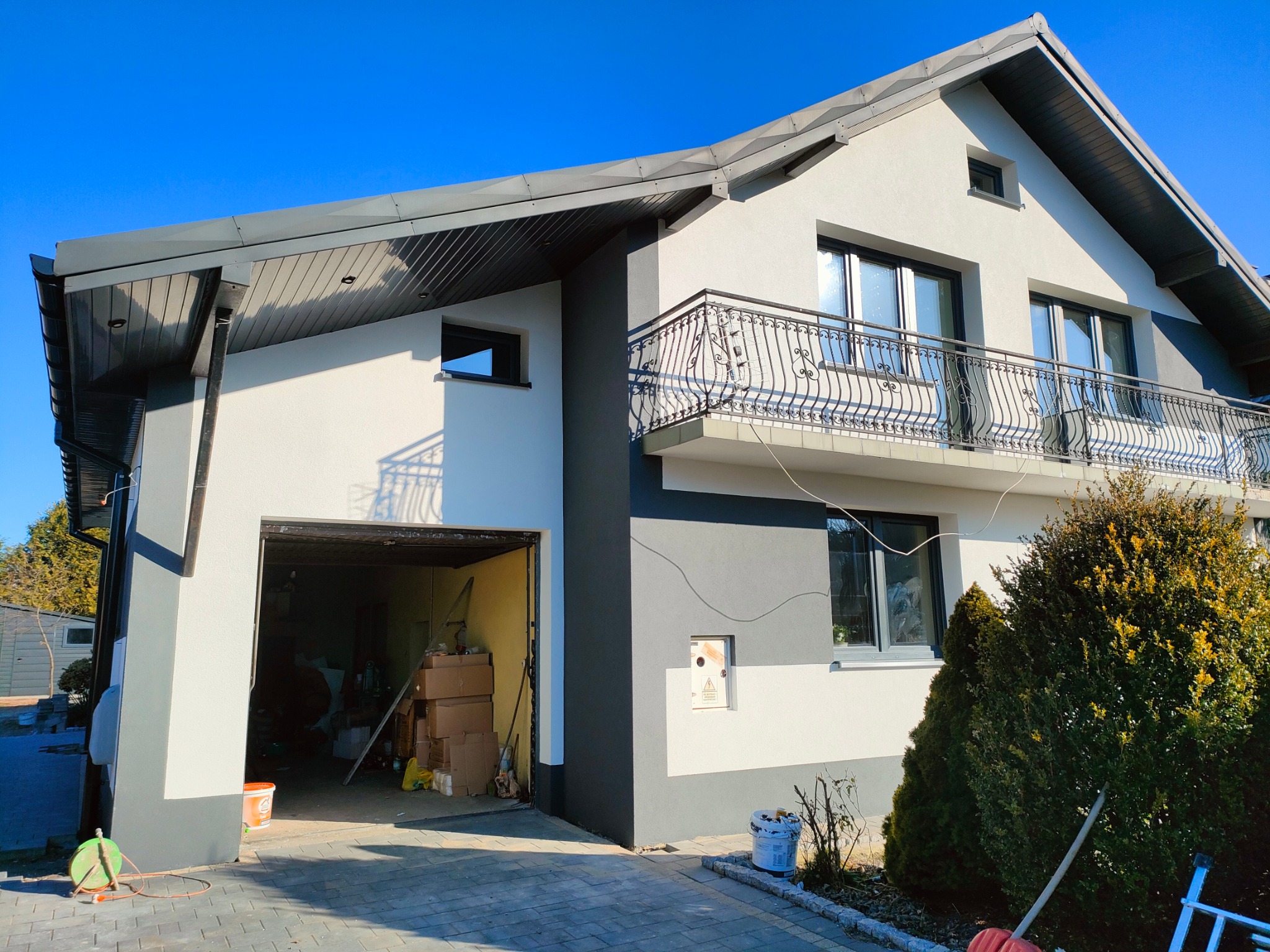 Dwukondygnacyjny dom z balkonem, świeżo pomalowaną elewacją w odcieniach szarości i bieli, widok z zewnątrz na otwarty garaż i podjazd z kostki brukowej.