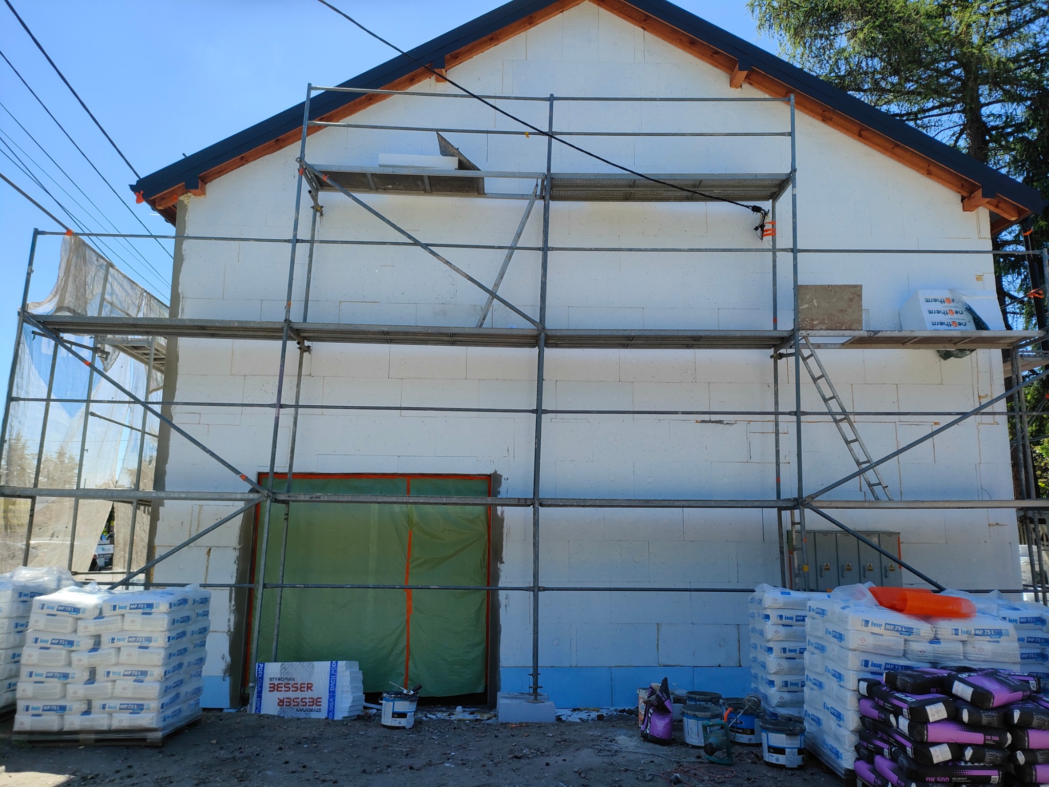 Budowa domu w trakcie ocieplania: białe ściany z pustaków, rusztowania, worki z zaprawą i materiały izolacyjne na placu budowy. Widoczne okna zakryte folią.