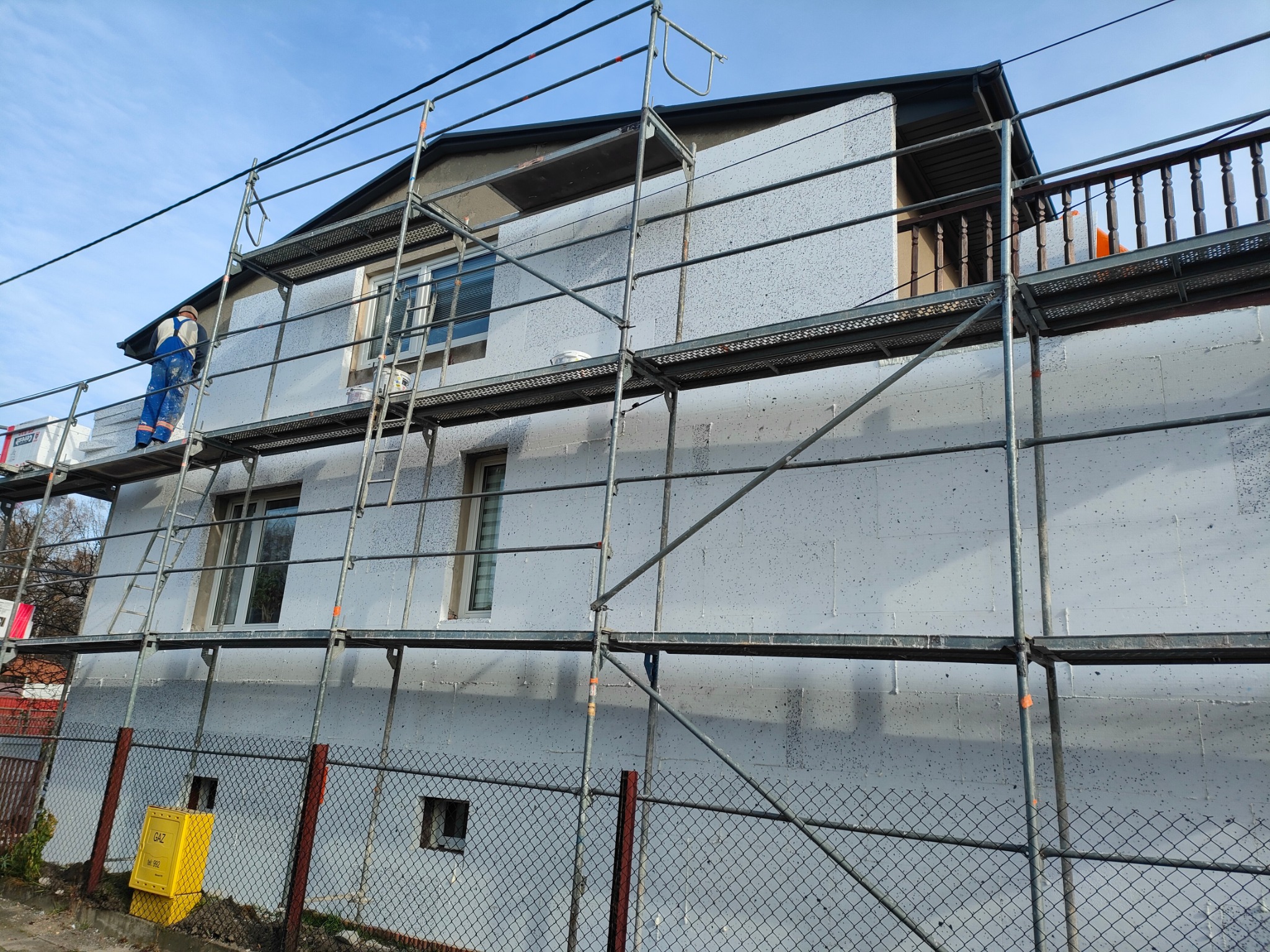 Dom w trakcie ocieplania, rusztowanie, pracownik w kombinezonie. Widok na ścianę pokrytą styropianem, przygotowaną do dalszych prac wykończeniowych.