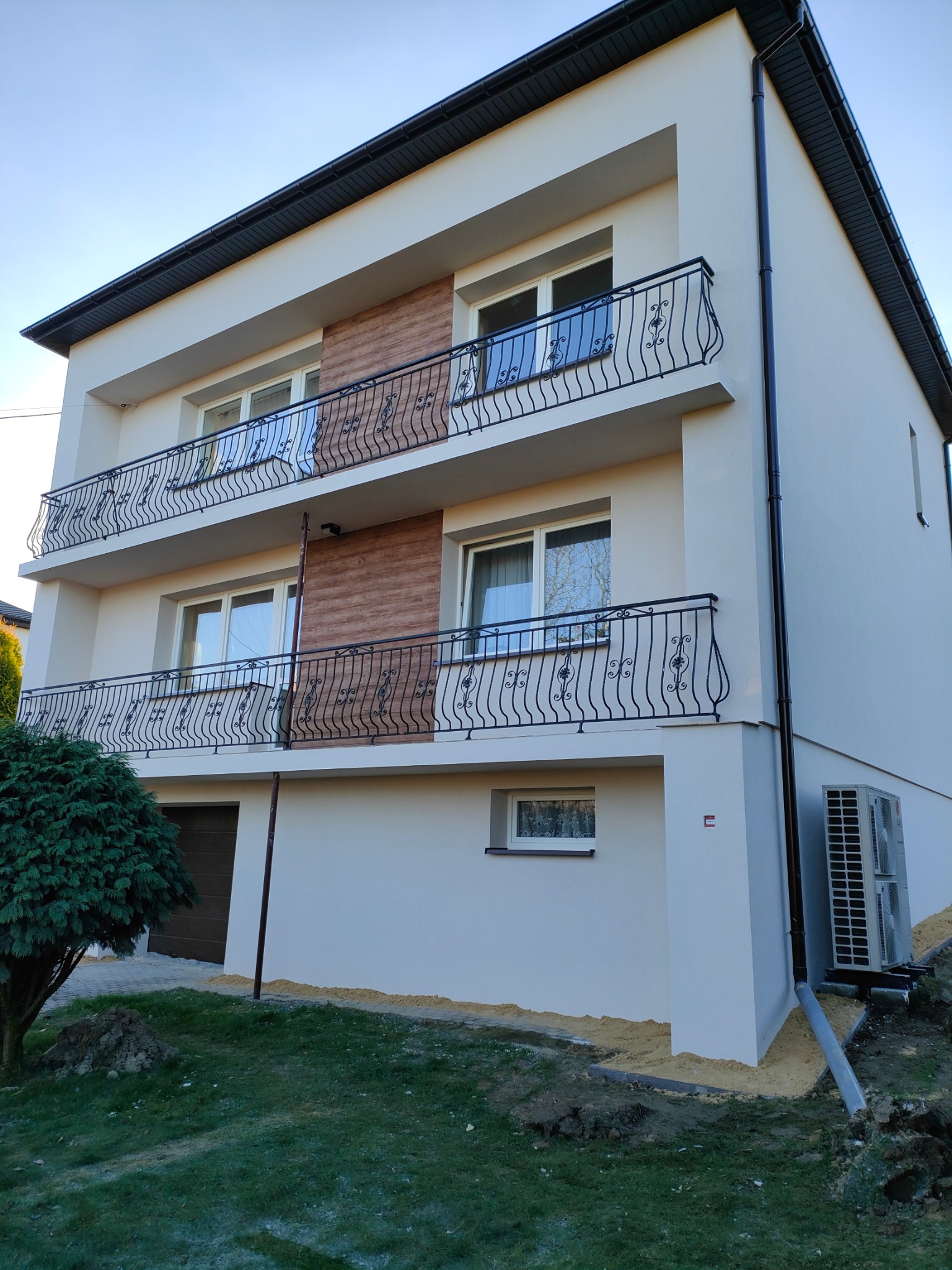 Dwupiętrowy dom z balkonami i elewacją w jasnym odcieniu. Kontrastują z nią czarne balustrady i rynny. Widoczny klimatyzator i fragment ogrodu z krzewem.