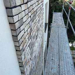 Usługi remonto budowlane - Elewacja domu z kamienia dekoracyjnego w odcieniach beżu i szarości, widoczna ze stojącego rusztowania. Detal wykończenia zewnętrznego budynku.