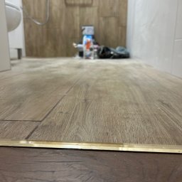 Usługi remonto budowlane - Ukończona podłoga w łazience z panelami imitującymi drewno, widoczna listwa progowa. W tle fragment toalety i ściany wyłożonej płytkami.