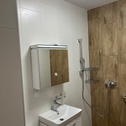 Usługi remonto budowlane - Nowoczesna łazienka z białymi płytkami i drewnopodobnymi panelami. Umywalka z szafką, lustro z oświetleniem, kabina prysznicowa z deszczownicą.