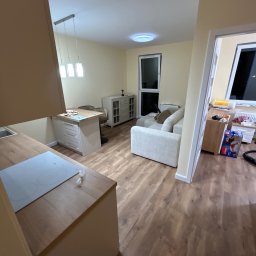 Usługi remonto budowlane - Jasne, przestronne wnętrze po remoncie: drewniana podłoga, aneks kuchenny z płytą indukcyjną, salon z sofą i oknem balkonowym. Nowoczesne oświetlenie sufitowe.