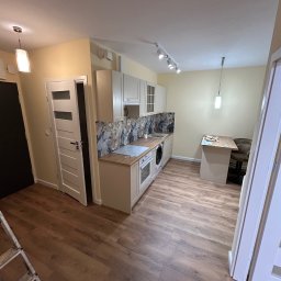 Usługi remonto budowlane - Jasna kuchnia po remoncie z drewnianą podłogą, wzorzystym panelem nad blatem i białymi szafkami. Widoczny stół z krzesłem i drabina malarska.