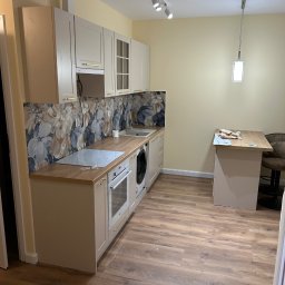 Usługi remonto budowlane - Jasna kuchnia po remoncie: beżowe szafki z drewnianym blatem, kwiatowy panel nad blatem, drewniana podłoga, oświetlenie punktowe i wisząca lampa.