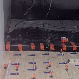 Usługi remonto budowlane - Układanie płytek podłogowych imitujących drewno z użyciem systemu poziomowania. W tle czarne płytki ścienne z marmurkowym wzorem i rolka taśmy.