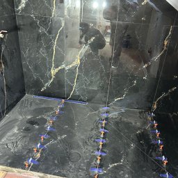 Usługi remonto budowlane - Układanie czarnych płytek imitujących marmur w kabinie prysznicowej. Widoczne niebieskie klipsy poziomujące. W tle odbicie glazurnika.