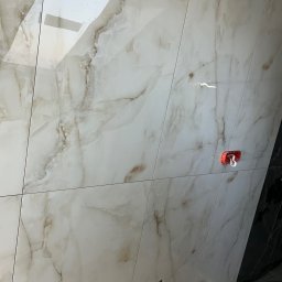 Usługi remonto budowlane - Ściana wyłożona błyszczącymi, beżowo-białymi płytkami imitującymi marmur. Widoczne otwory na armaturę i uchwyt na słuchawkę prysznicową. Fragment czarnej ściany w tle.