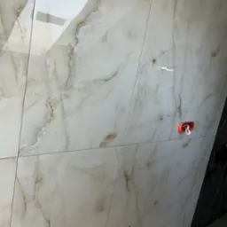 Ściana wyłożona błyszczącymi, beżowo-białymi płytkami imitującymi marmur. Widoczne otwory na armaturę i uchwyt na słuchawkę prysznicową. Fragment czarnej ściany w tle.