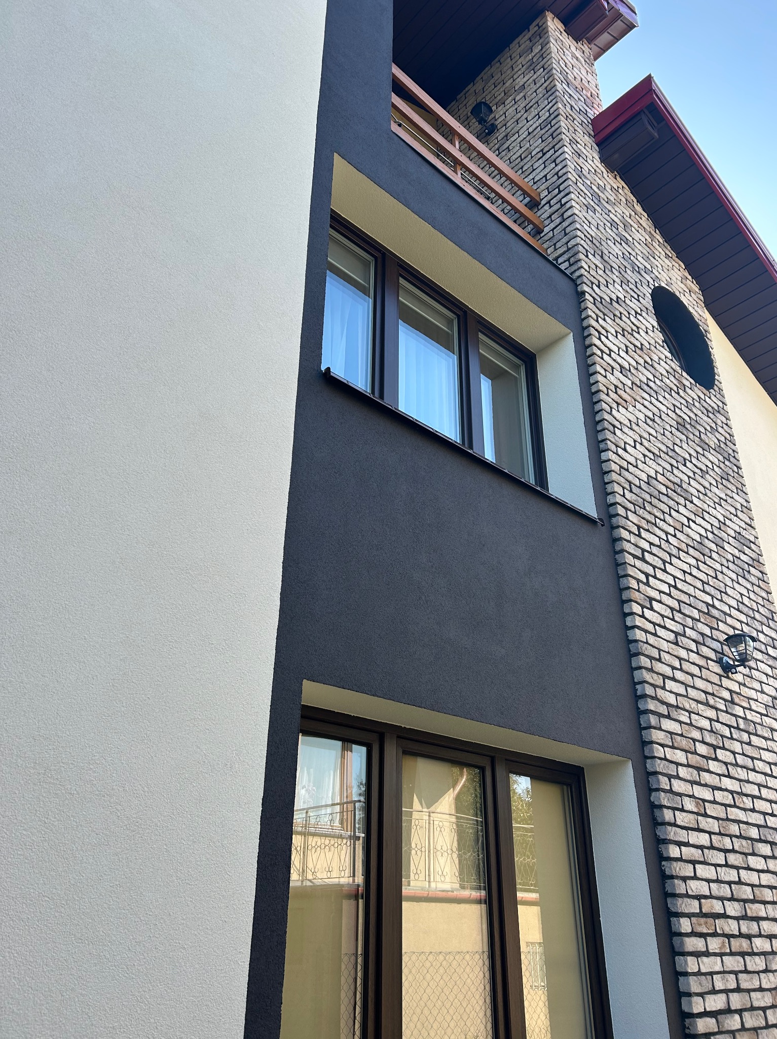 Elewacja domu z fragmentem balkonu, łącząca tynk w odcieniach bieli i szarości z elementami z cegły. Widoczne okna z brązowymi ramami i fragment dachu.