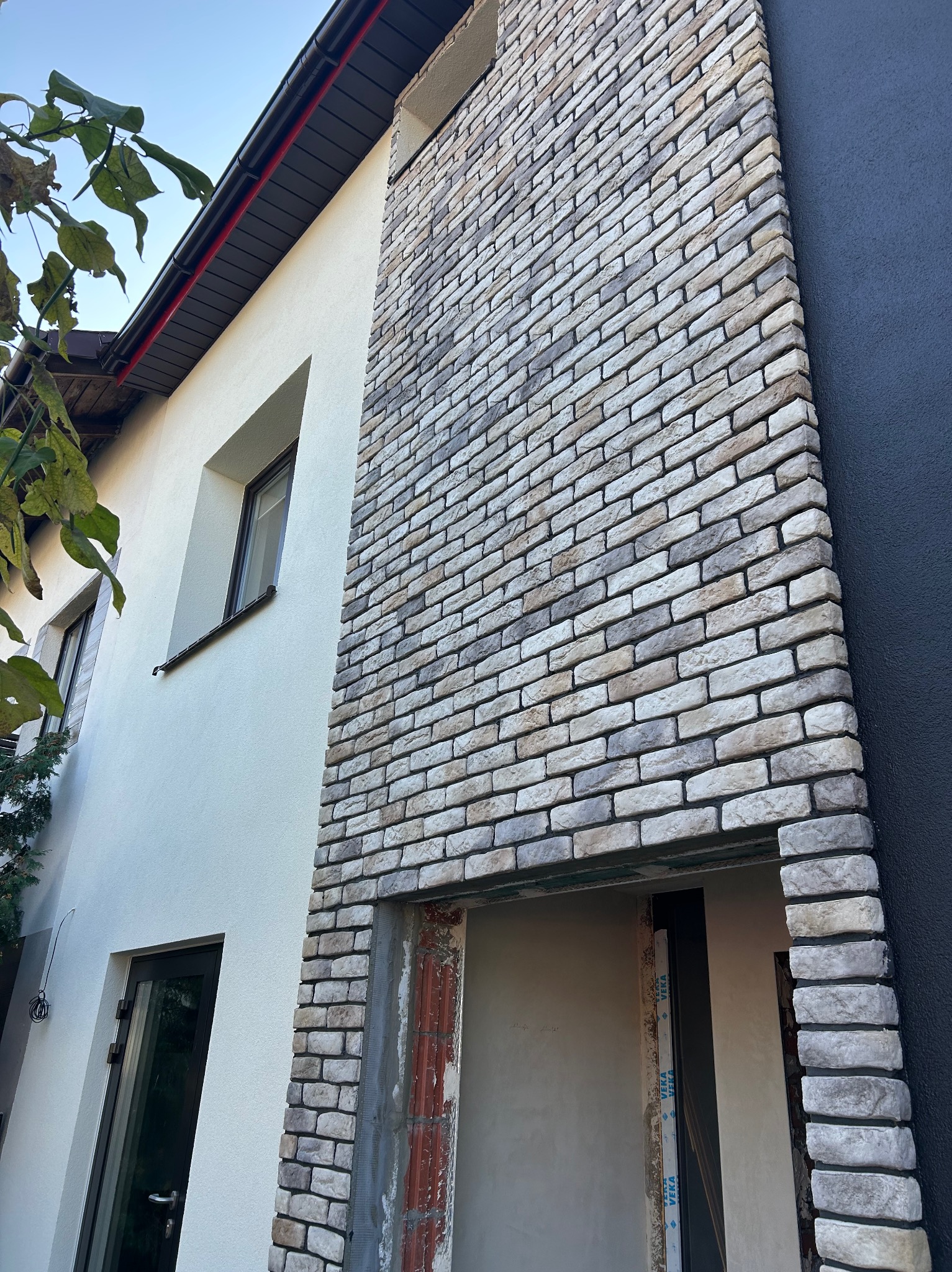 Elewacja domu z dekoracyjną okładziną z cegły w odcieniach szarości i beżu, kontrastująca z białą i grafitową ścianą. Widoczne okno i fragment drzwi.